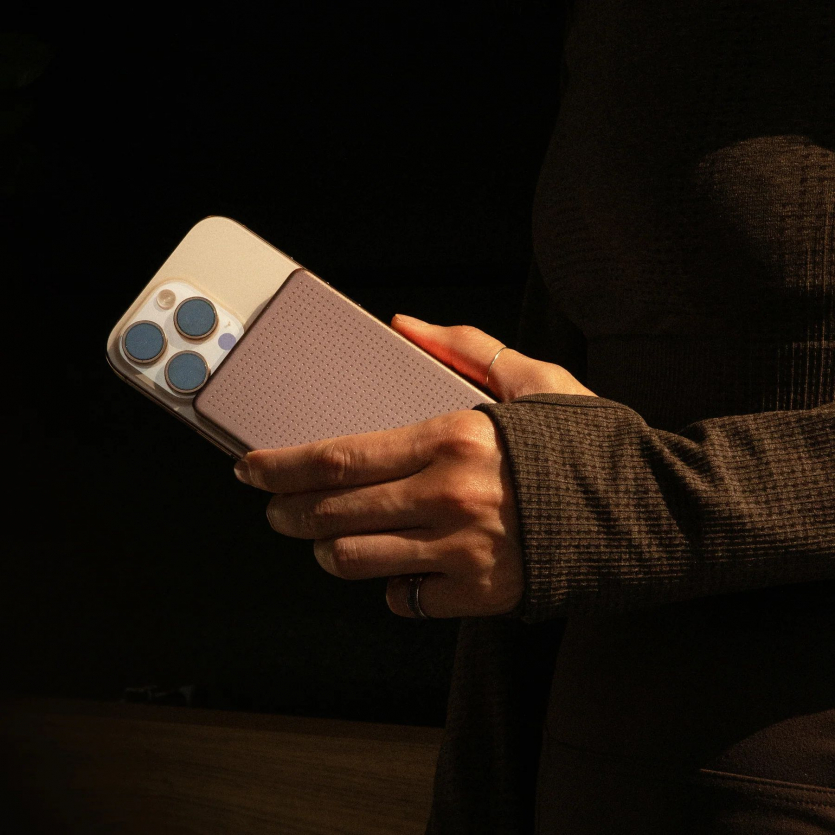 Рука тримає золотистий iPhone Pro під час заряджання від прикріпленого рожевого павербанка Native Union Active 5000 mAh.