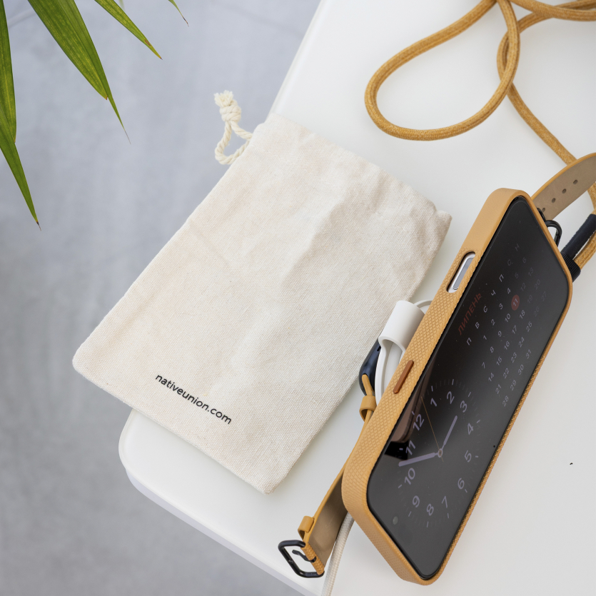 Смартфон iPhone у фірмовому чохлі з ремінцем, кабель та тканинний мішечок Native Union на білому столі.