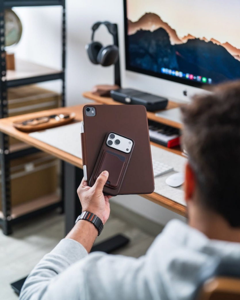 Чоловік тримає iPad та iPhone у шкіряних чохлах Nomad Horween на фоні стильного робочого місця з монітором та аксесуарами.